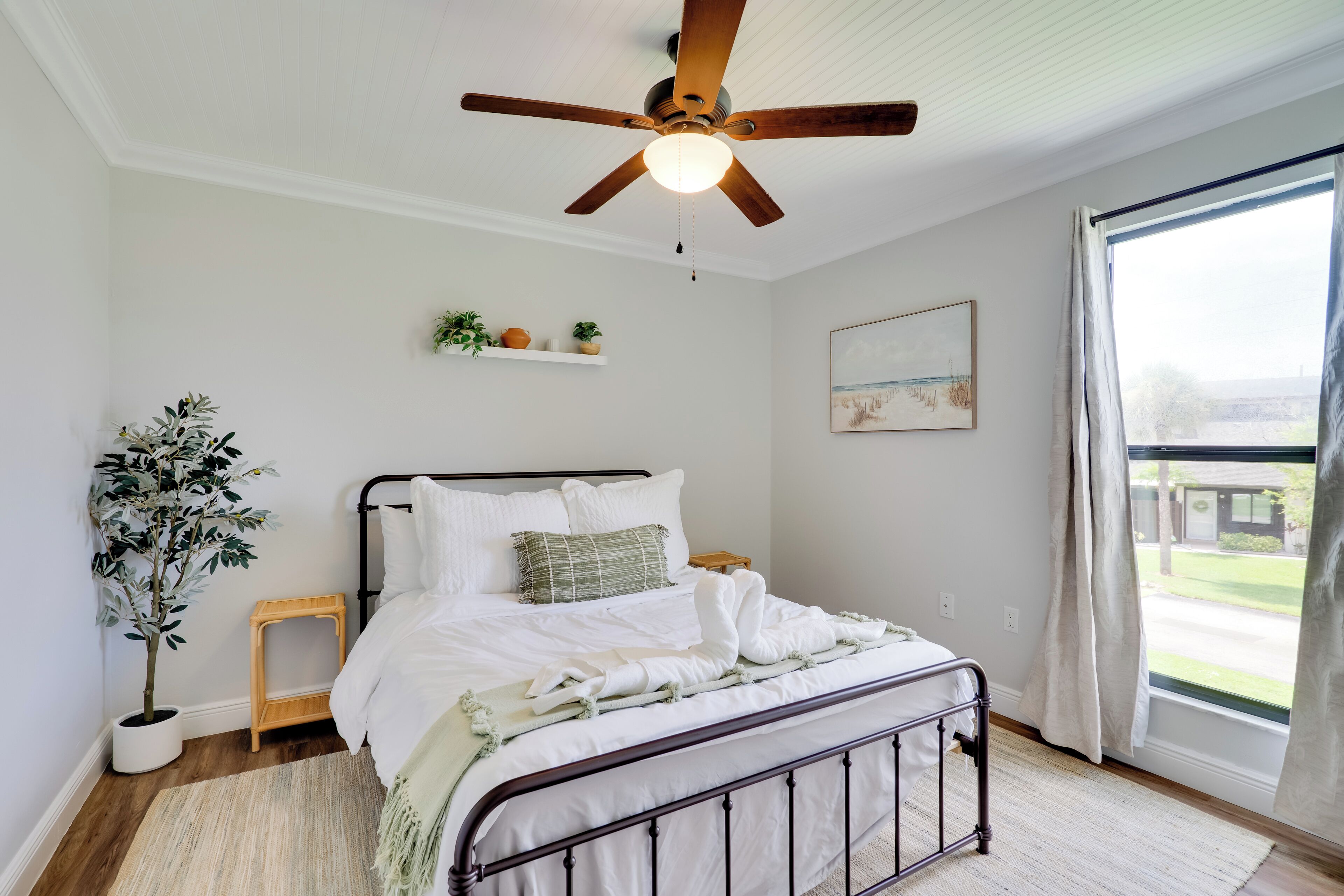 A bedroom with a bed, ceiling fan, potted plants, and a window with curtains.