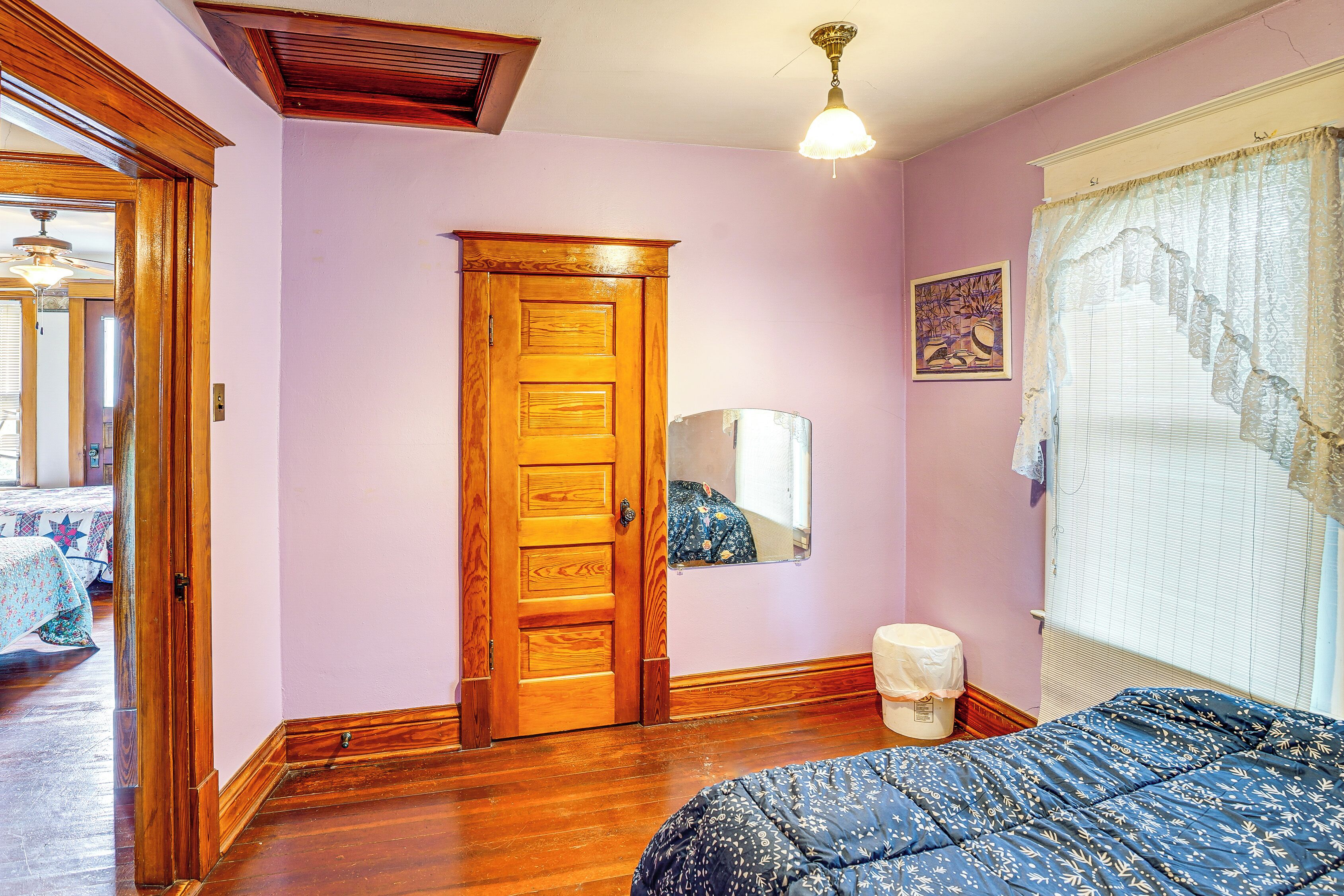 Una habitación con puerta de madera, una cama con colcha estampada, una ventana con cortinas de encaje y una lámpara en el techo.