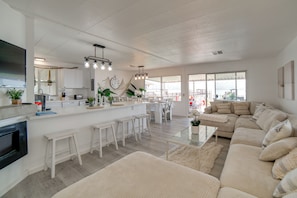 A spacious living area with a large beige sofa, a glass coffee table, and a kitchen area with white cabinetry and a central island. - Hot Tub, Views & Dock! Playful Home on Clear Lake (Lakeport)