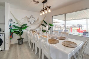 A dining area with a table set for six, white chairs, a large window with a view of the sea, a clock on the wall, and a hanging plant. - Hot Tub, Views & Dock! Playful Home on Clear Lake (Lakeport)