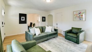 A living room with a sectional sofa, a green armchair, and a gray rug. There are framed pictures on the walls and a door in the background. - Walk to Ritter Park: Renovated Home Near Marshall! (Huntington)