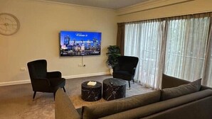 Una sala de estar con sofá, dos sillones, una mesa de centro, televisor y un ventanal grande con cortinas.