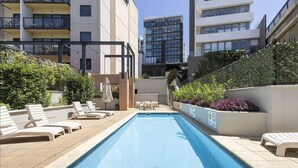 Una piscina con sillones de descanso, rodeada de edificios con balcones y un seto.