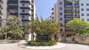 Un edificio residencial de varios pisos con balcones, un área ajardinada con árboles y un letrero que indica el nombre 'Mounts Bay Village'.