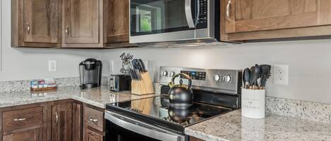 A kitchen with wooden cabinets, a stainless steel microwave, a coffee maker, a kettle, and a utensil holder.