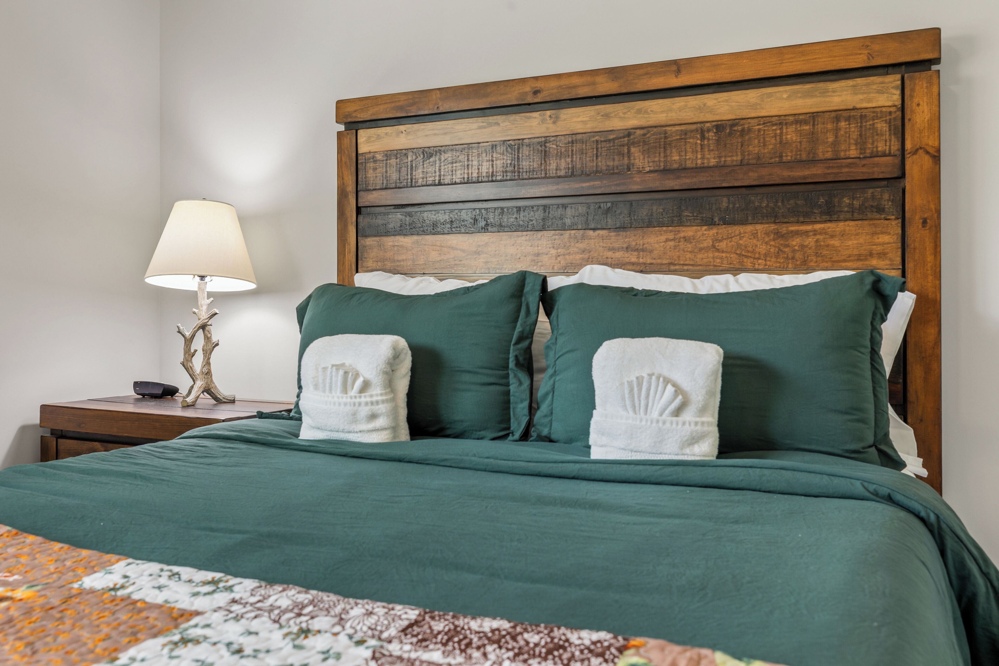 A neatly made bed with green bedding and two white towels, a bedside lamp, and a wooden headboard.