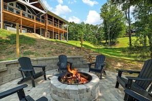 A stone fire pit with chairs around it, set on a patio with a view of a building and a grassy hill. - Cozy Blue Ridge Cabin: Game Room & Mountain Views (Blairsville)