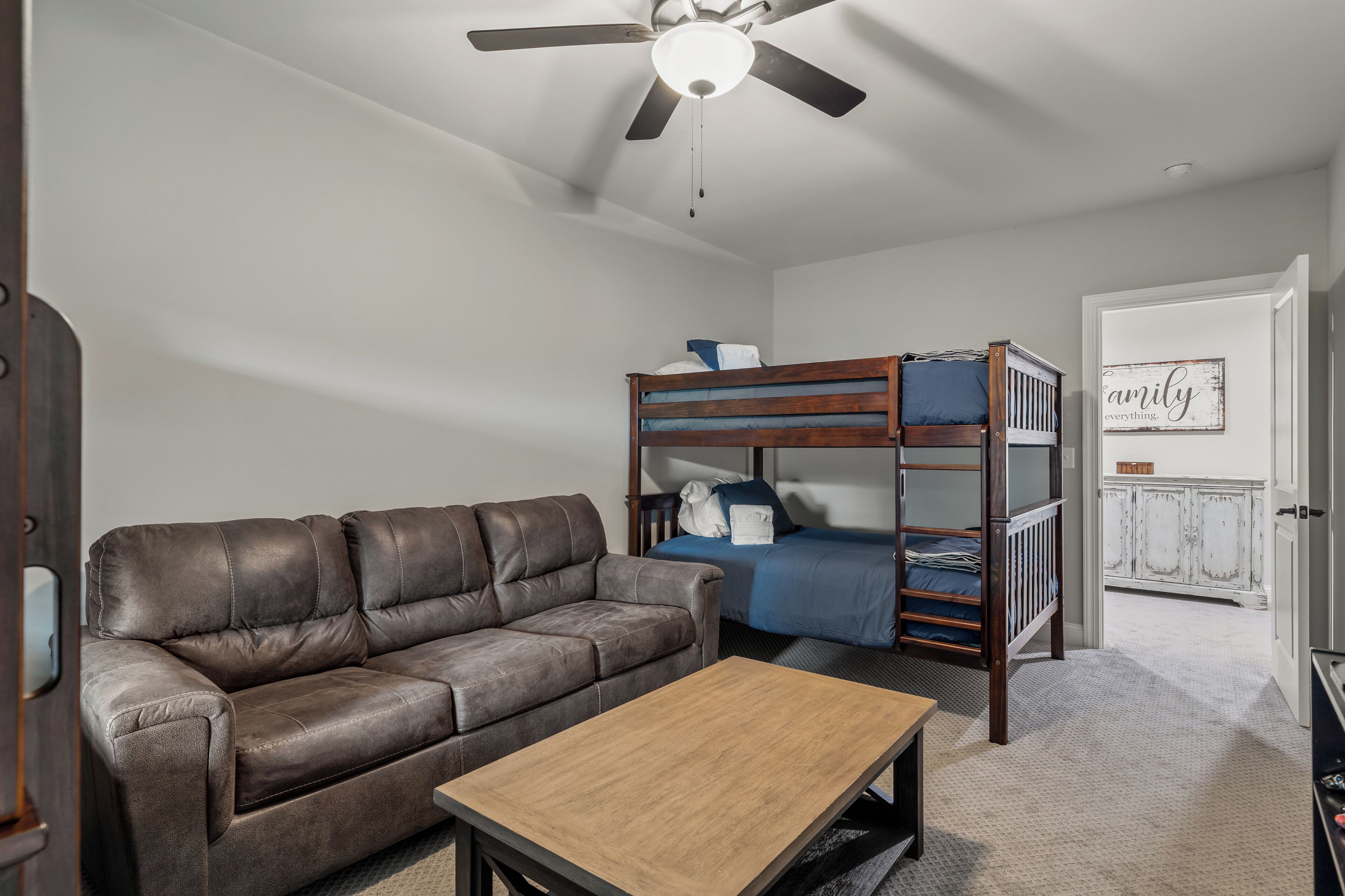 A room with a brown leather sofa, a wooden coffee table, a bunk bed with blue bedding, and a ceiling fan.