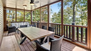 Una terraza techada al aire libre con una mesa y sillas de madera, un sofá, y vistas a árboles y una masa de agua.