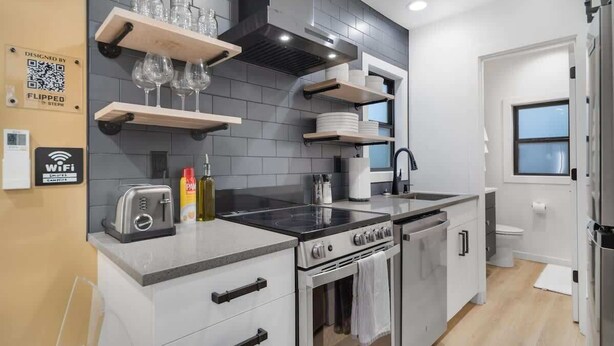 A modern kitchen with grey tiles, stainless steel appliances, and wooden shelves.