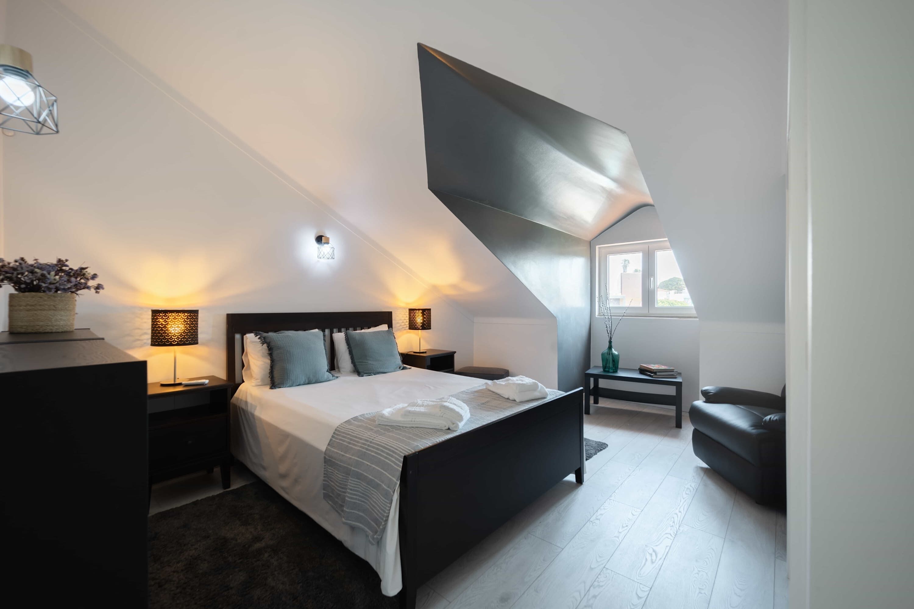 A modern bedroom with a large bed, bedside tables with lamps, a dark sofa, and a window with natural light.