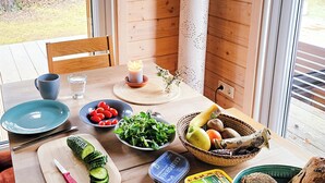 Una mesa de madera con diversos alimentos, utensilios, una vela y una ventana que ofrece vistas a una casa y árboles.