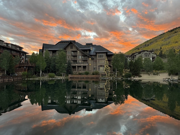 Un hotel de varios pisos con su reflejo en un lago tranquilo, rodeado de árboles y un paisaje montañoso.