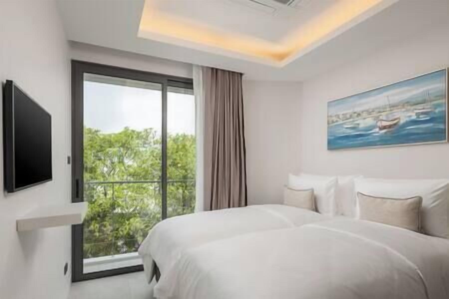 A hotel room with a large bed, a flat-screen TV mounted on the wall, a painting of a coastal scene, and a view of greenery through the window.