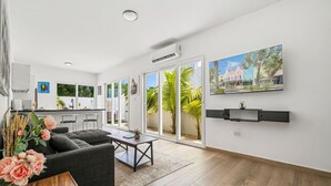 Un salon moderne avec un canapé gris, une table basse et un vase de fleurs. Une porte-fenêtre coulissante donne accès à un balcon d’où l’on peut admirer des palmiers et un immeuble.