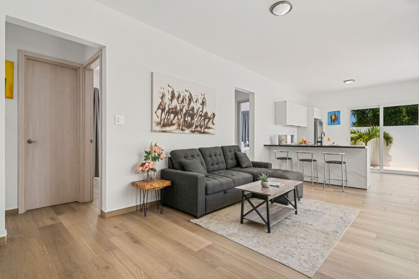 Een moderne woonkamer met een grijze bank, een salontafel en een schilderij van paarden. Er is een keukenhoek met een bar en stoelen.