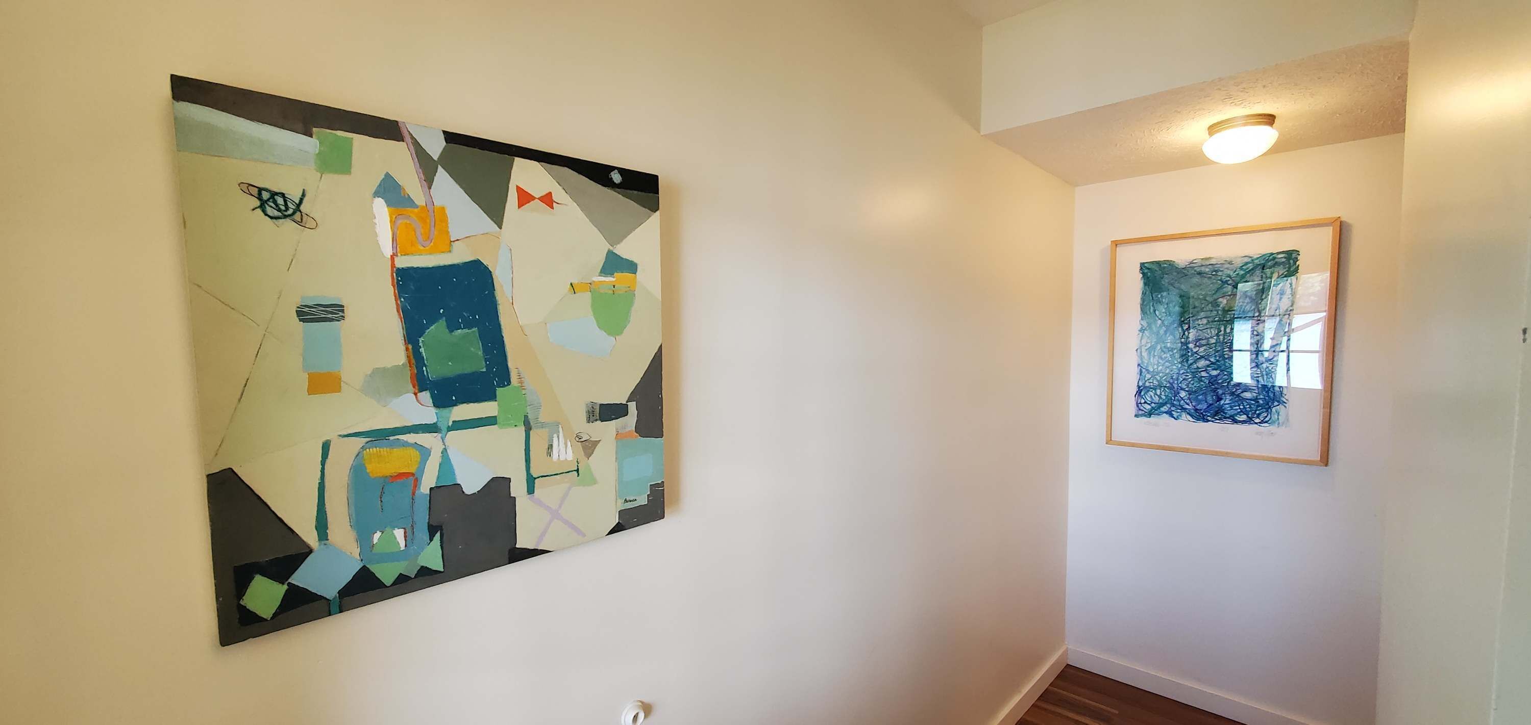 Un couloir avec deux œuvres d’art accrochées aux murs, un luminaire au plafond et un plancher en bois.
