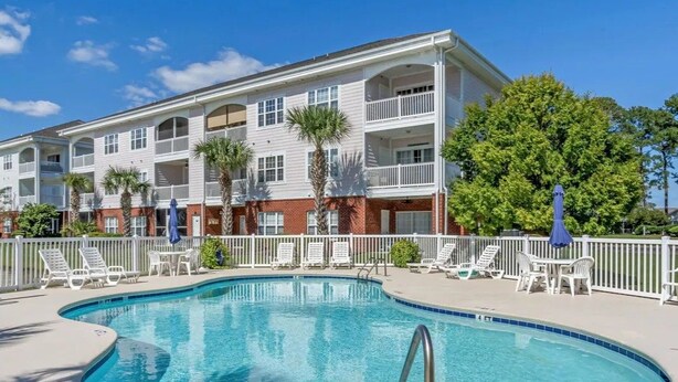 A pool area with lounge chairs and a building with multiple balconies. - Pet Friendly 2BR Condo, Spacious w/ Vaulted Ceilings! (Little River)