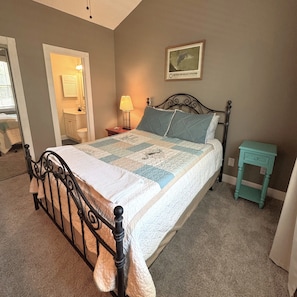 A bedroom with a bed, a nightstand, a lamp, a small table, a framed picture on the wall, and a door leading to another room.