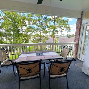 A covered patio with a table and chairs, a white railing, and a view of trees and houses. - Pet Friendly 2BR Condo, Spacious w/ Vaulted Ceilings! (Little River)