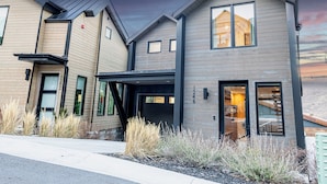 A modern two-story house with a dark roof, large windows, and a visible staircase leading to the upper floor. - Modern Old Town Home by InvitedHome | Ski-In/Out, Walk to Town, Hot Tub (Park City)
