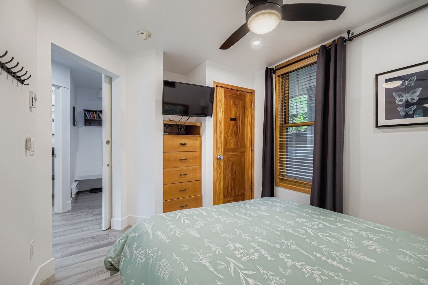 A bedroom with a bed, a ceiling fan, a window with curtains, and a door leading to another room.