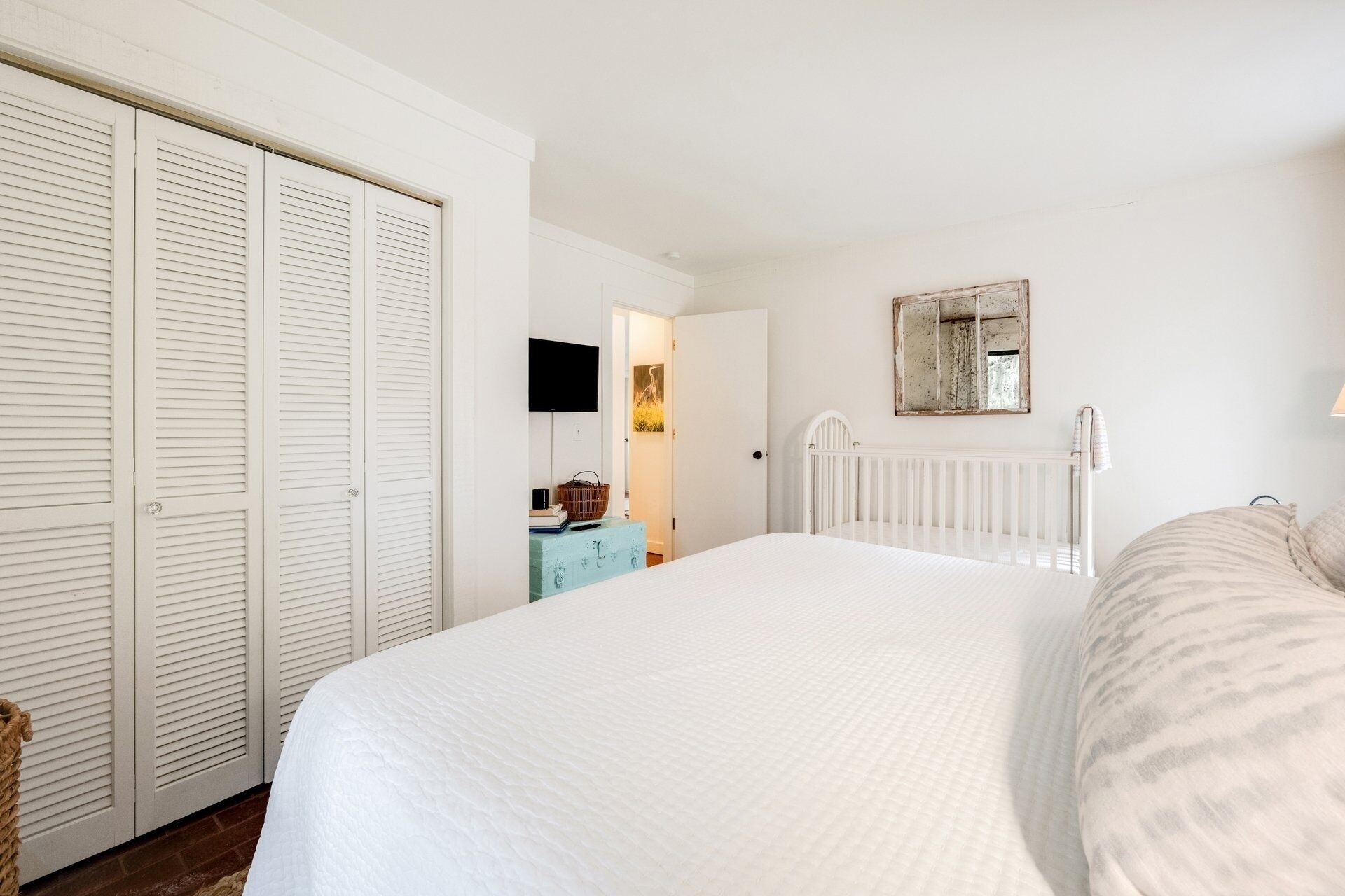 A bedroom with a large bed, a white wardrobe, a crib, and a wall-mounted TV.
