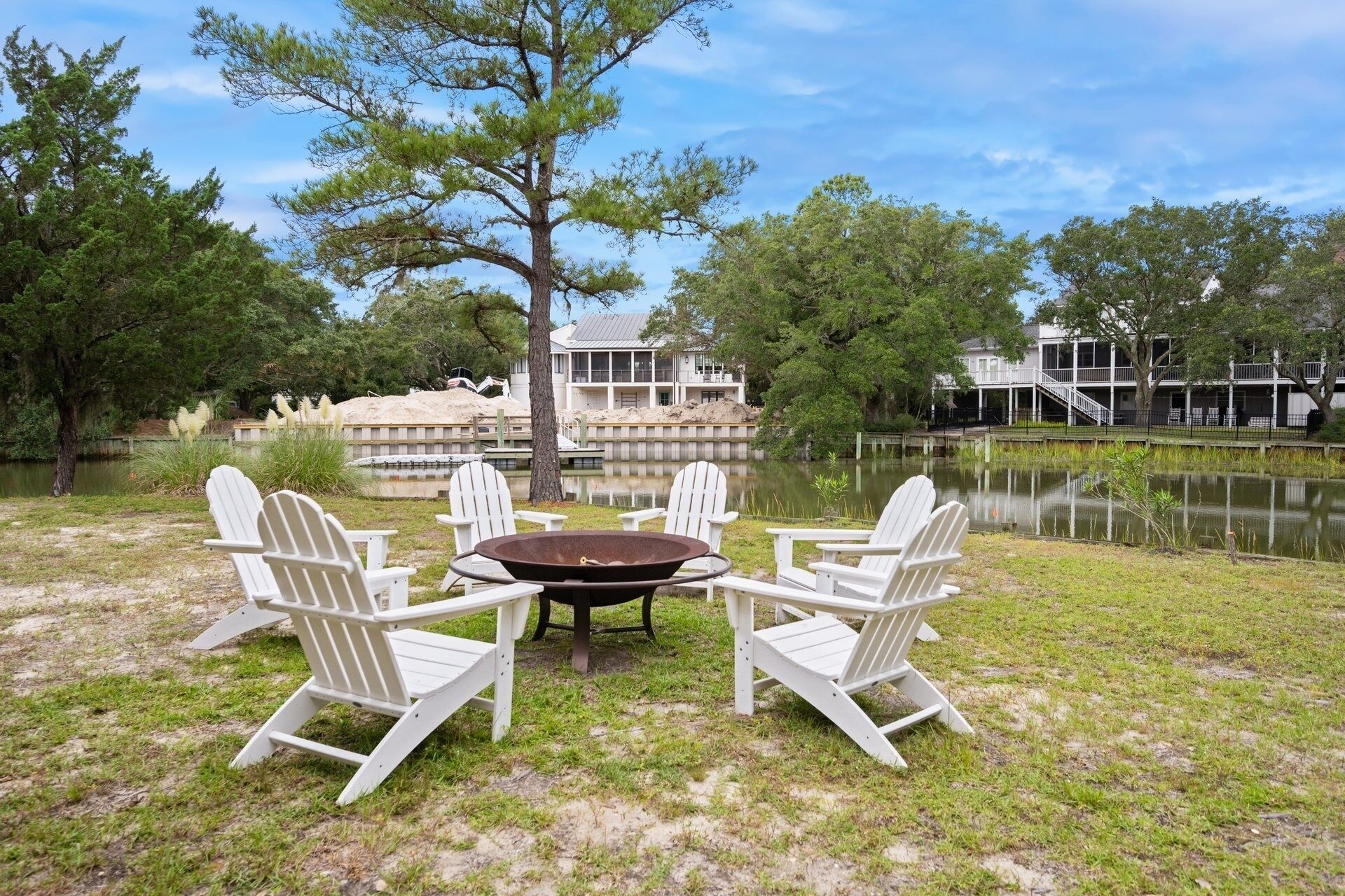 A lakeside outdoor seating area with white Adirondack chairs and a round table, surrounded by a grassy lawn and trees.