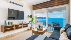 Una sala de estar moderna con televisor de pantalla plana, estanterías de madera, una mesa de centro y un sofá. Un amplio ventanal corredera da a un balcón con vistas al mar y al paisaje urbano.