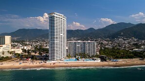 Un paisaje urbano costero con un imponente hotel de gran altura que cuenta con varios balcones, una playa con sombrillas y montañas al fondo.