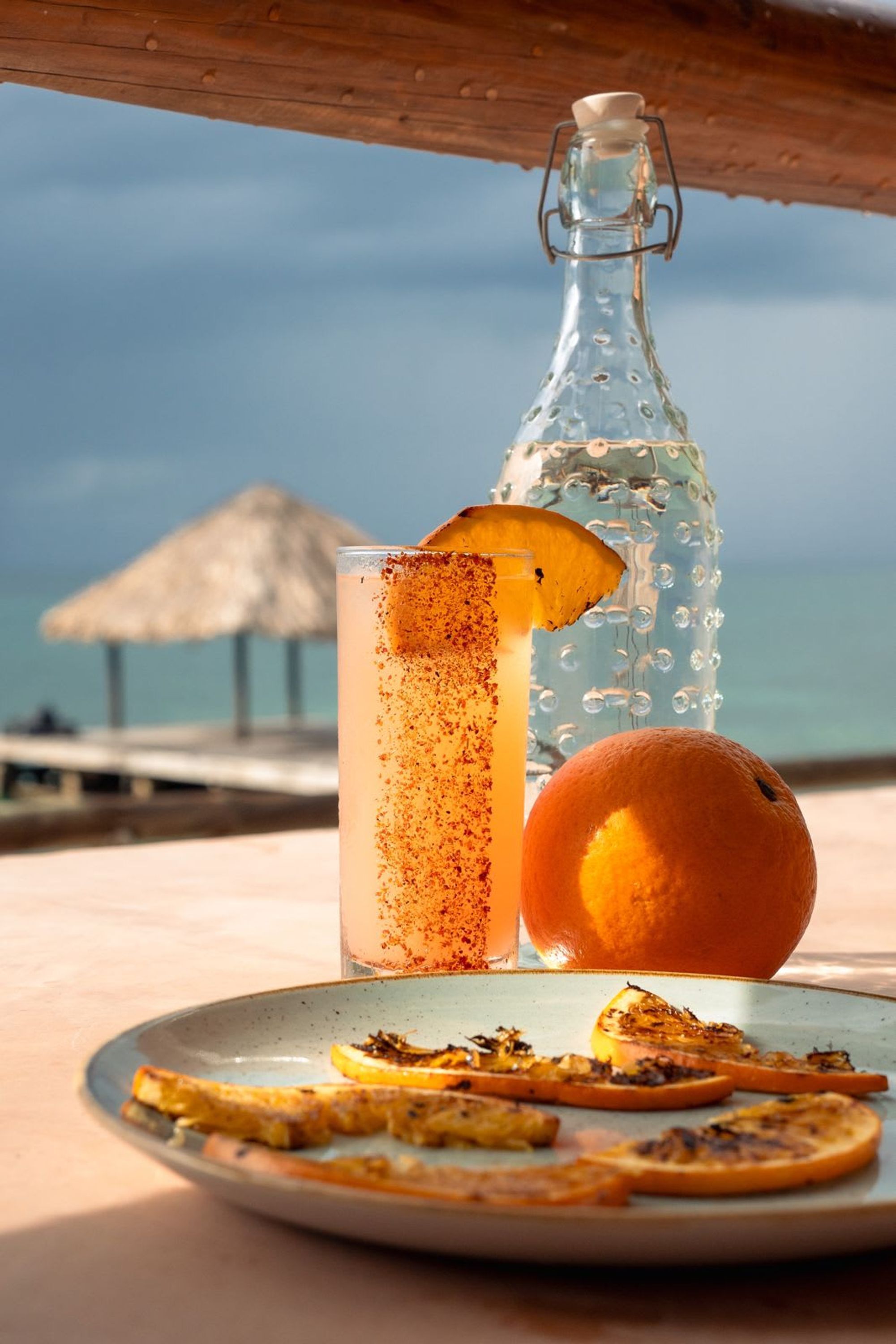 A glass of cocktail with a slice of orange and a cinnamon sprinkle, a bottle of water, and a whole orange on a plate with toasted bread slices.