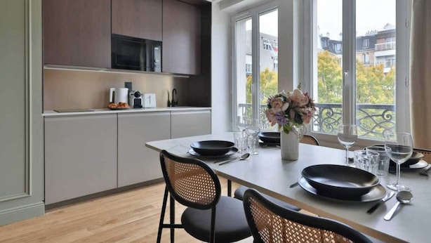 A modern kitchen with dark cabinets, a dining table set for two, and a view of buildings through large windows. - Modern Apartment - 1br/4p - Canal St-martin (Paris)