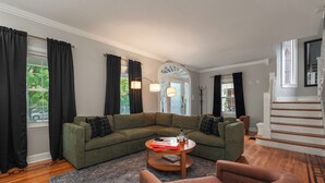 A living room with a green sectional sofa, a round coffee table, and a brown armchair. There is a staircase to the right and a door with a stained glass window. - The Reserve ~ 5BR Fun! Hot Tub, Fire Pit, Deck, Walkable To Bardstown Rd (Louisville)