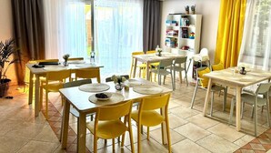 Un coin repas avec des chaises et des tables jaunes, une bibliothèque et une fenêtre avec des rideaux.