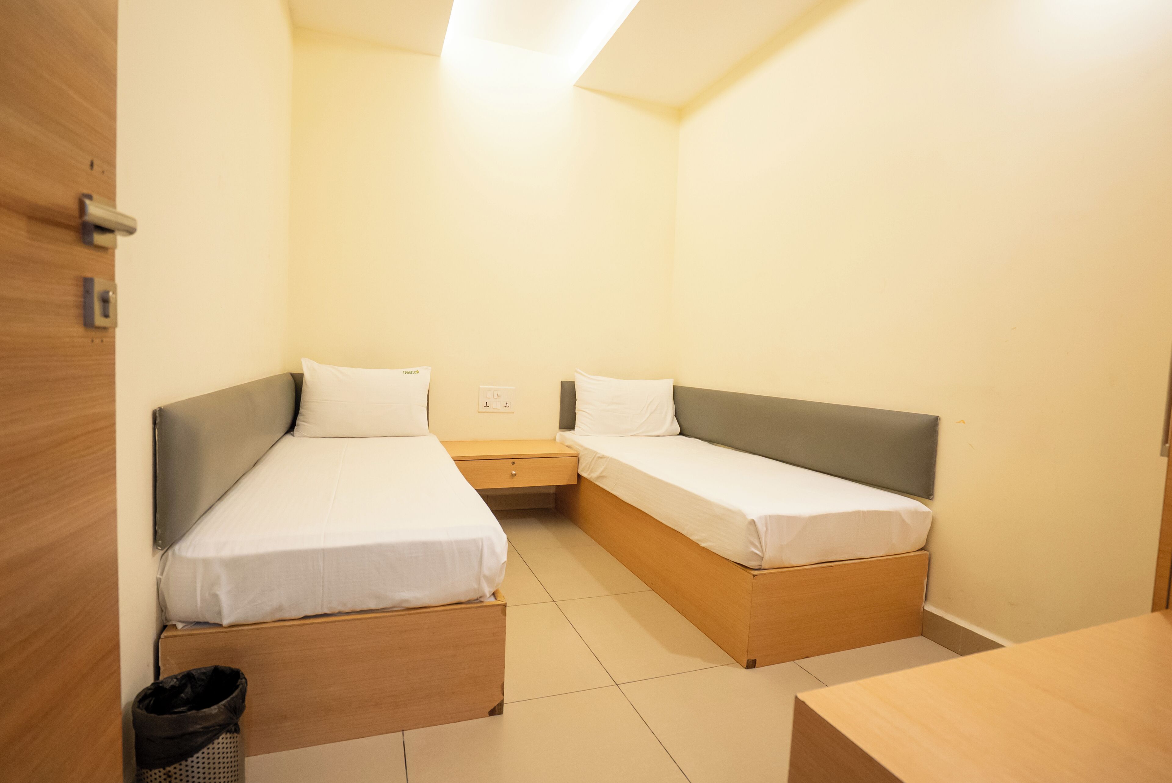 A small, clean room with two single beds, a wooden nightstand, a trash bin, and a wooden desk.