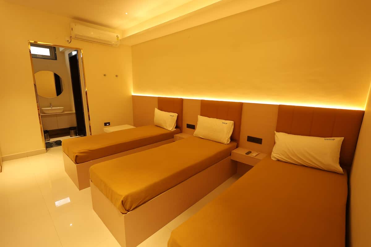 Habitación de hotel con tres camas individuales, una mesita de noche con lámpara y baño con lavabo y espejo.
