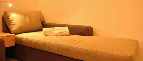 Una habitación pequeña con una cama individual, un sofá cama marrón, una mesita de noche con una lámpara y dos toallas con la inscripción 'fresh up'.