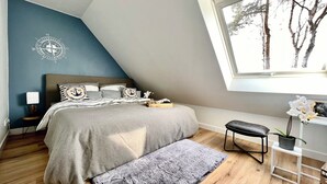 A bedroom with a sloped ceiling, a bed with a grey bedspread, a bedside table with a lamp, a small stool, and a window with a view of trees.