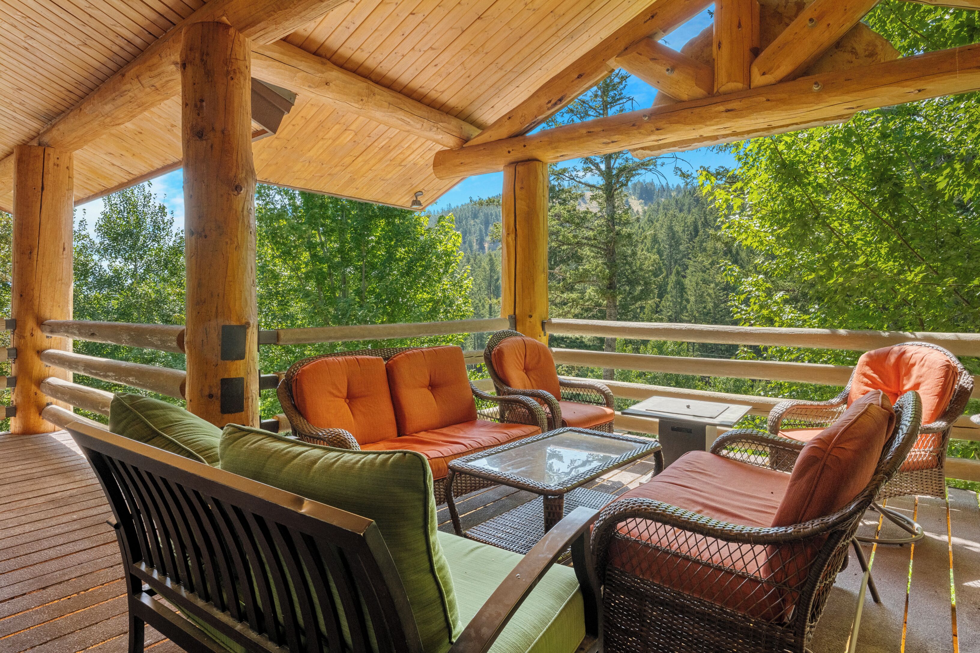 Une terrasse en bois, dotée de meubles en osier et d’une table en verre, qui offre une vue sur une zone boisée.