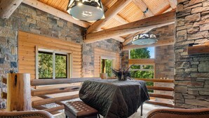 Interior de una cabaña de troncos con pared de piedra, vigas de madera, una mesa cubierta, sillones de cuero y ventanas amplias.