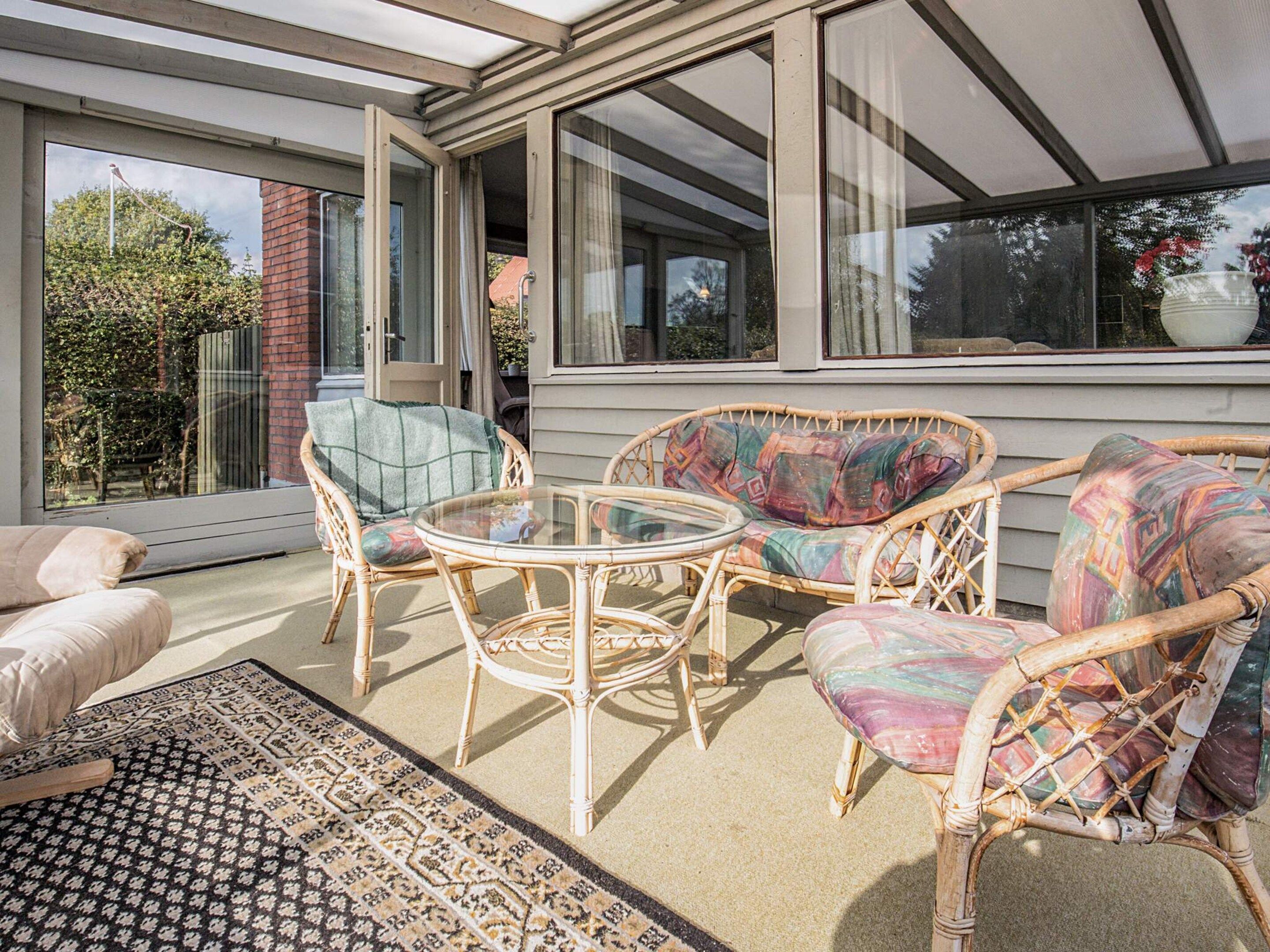 A sunlit patio with wicker furniture, a glass table, and a patterned rug.