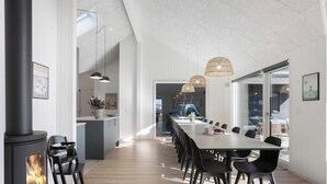 Un interior moderno con una larga mesa de comedor, sillas negras, una isla central con pileta, luces colgantes y una chimenea a la vista.