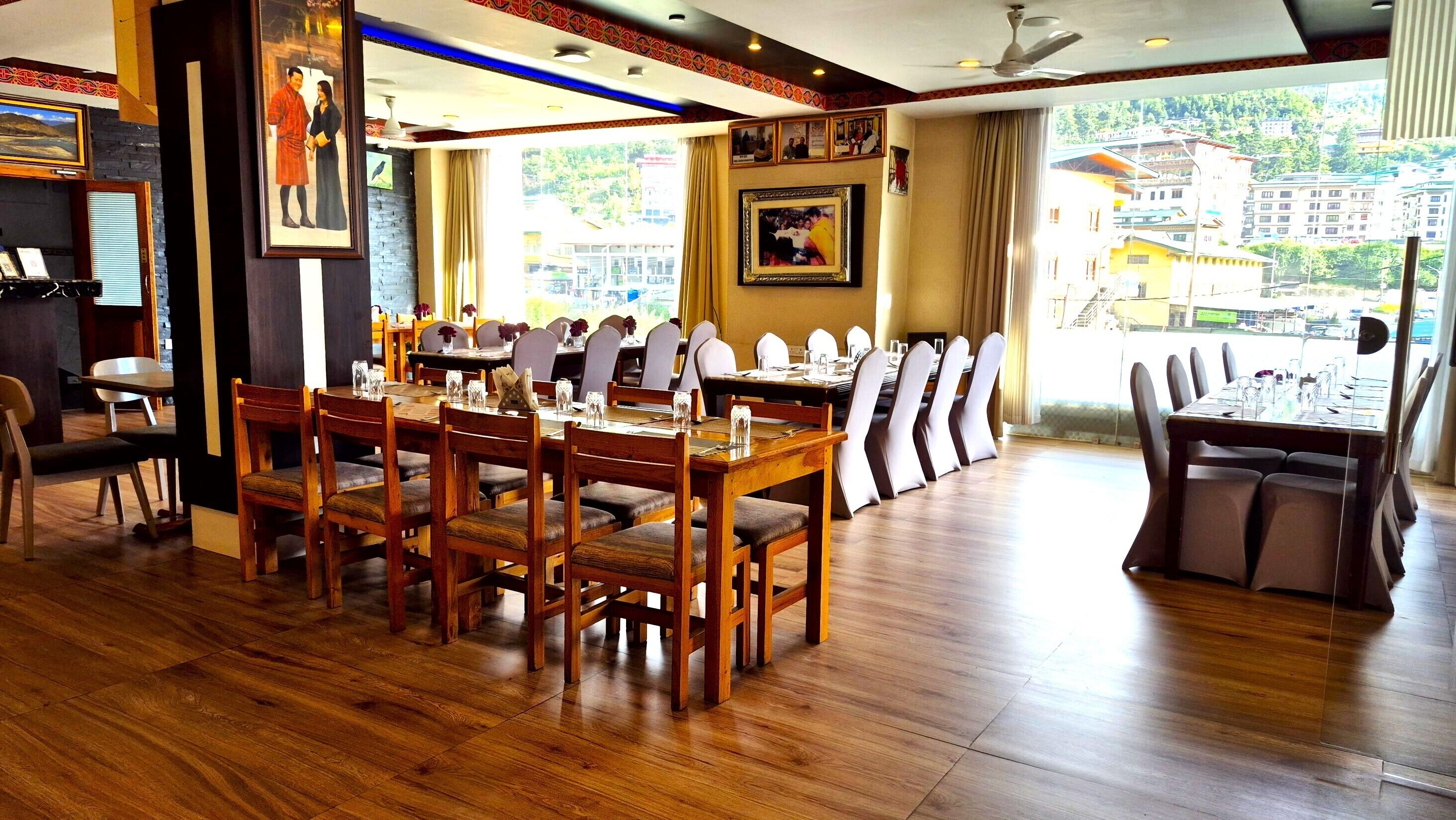 A dining area with wooden tables and chairs, a large window with a city view, and framed pictures on the walls.