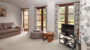 A living room with a sofa, a bean bag, a TV stand, and large windows with curtains. - The Milking Shippon at Thorne (Torrington)