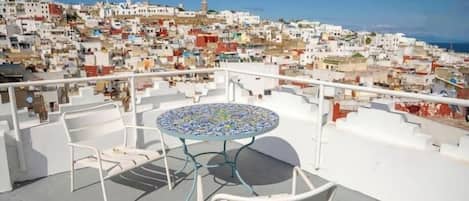 A rooftop terrace with a round table and two chairs, overlooking a densely packed coastal town with white buildings and a clear blue sky.