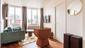 Een moderne woonkamer met een bank, een rond poefje, een televisietoestel en een groot raam met gordijnen.