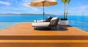 Una terraza de madera con dos sillones reclinables y una sombrilla grande, con vista a un mar azul y cristalino.