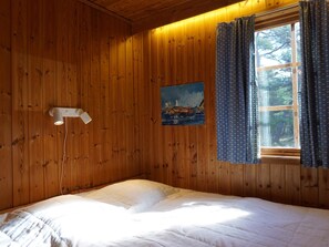 Huone, jonka seinät ovat puuta, valkoisilla lakanoilla varustettu sänky, seinällä oleva taulu ja ikkunassa olevat verhot.