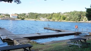 Puinen laituri, joka ulottuu tyynen vesialueen päälle. Oikealla puolella on piknikpöytiä ja kyltti, jossa on kuva pelastusrenkaasta.