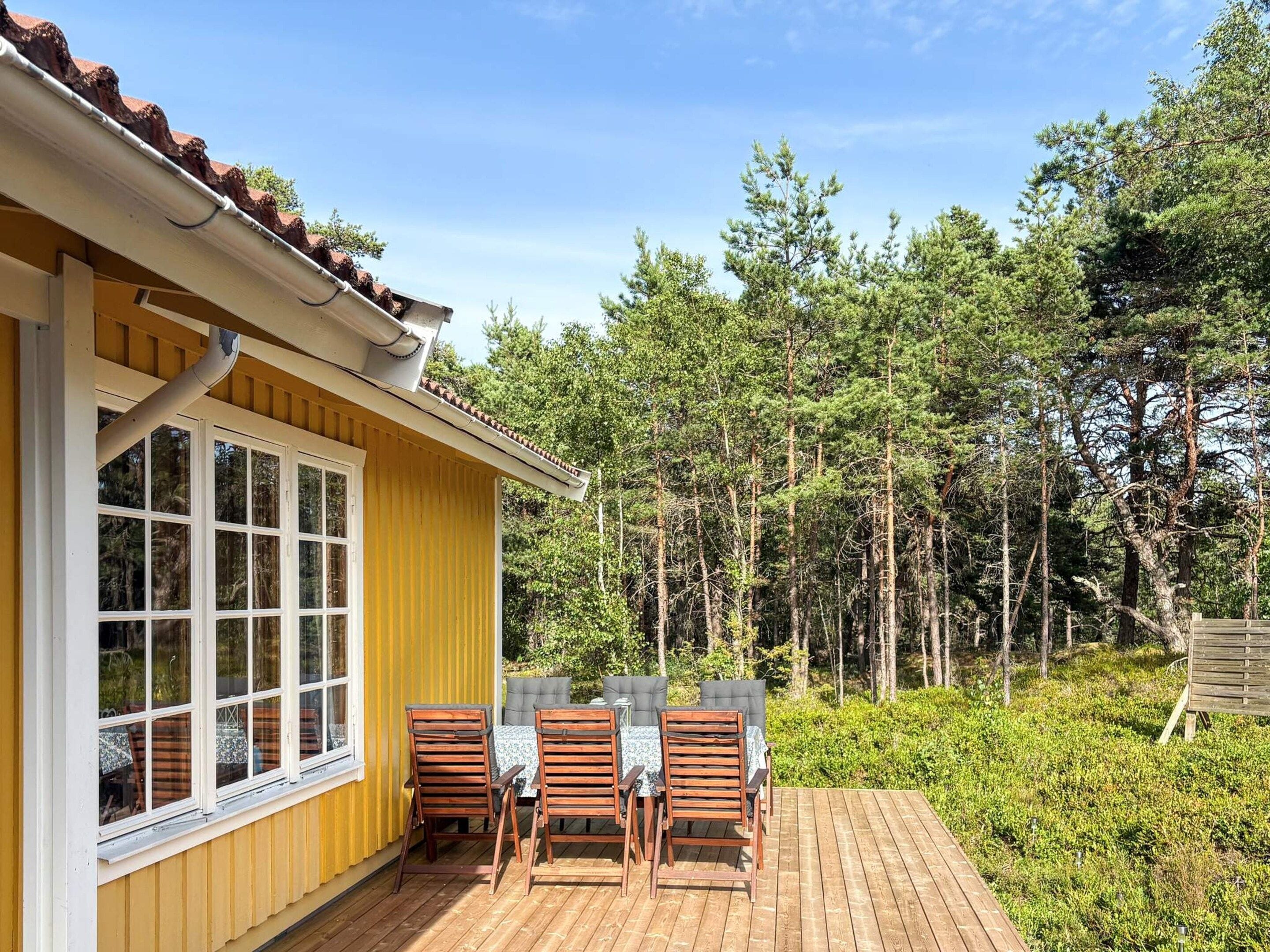Un bâtiment jaune avec une terrasse, une table entourée de chaises et une forêt en arrière-plan.